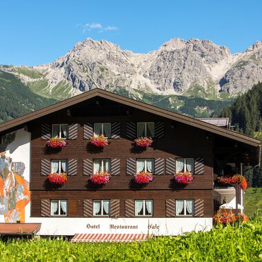A charming chalet in the mountains with flower balconies. In the background, impressive mountain peaks and a clear sky can be seen. | © Hotel Alte Krone
