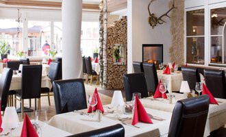 Ein elegantes Restaurant mit modernen Tischen und Stühlen. Die Tischdecken sind cremefarben und die Servietten in Rot, während ein Kamin im Hintergrund für eine gemütliche Atmosphäre sorgt. | © Aparthotel Kleinwalsertal Haller GmbH & Co KG