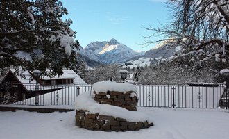 Eine winterliche Landschaft mit schneebedeckten Bergen im Hintergrund. Der Vordergrund zeigt einen steinernen Kamin und verschneite Dächer. | © Hotel Bellevue | I. Malouvier