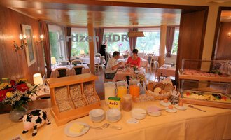 Ein einladender Frühstücksraum mit einem Buffet voller Speisen und Getränken. Im Hintergrund sitzen zwei Personen am Tisch und genießen ihr Frühstück. | © Hotel Bellevue | Werbewind GmbH
