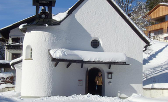 Eine kleine weiße Kapelle im Schnee mit einem Glockenturm. Im Hintergrund sind schneebedeckte Bäume und ein Holzgebäude sichtbar. | © Hotel Bellevue | I. Malouvier