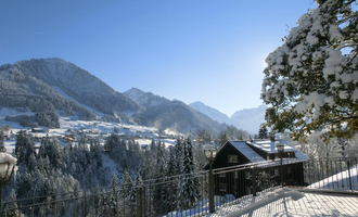 A winter landscape with snow-covered mountains and trees. In the foreground, there is a wooden house with a balcony overlooking the mountains. | © Hotel Bellevue | I. Malouvier