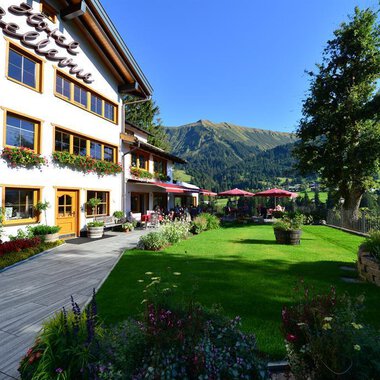 A beautifully designed house with balconies and blooming flowers. In the background, green mountains and a clear sky can be seen. | © Hotel Bellevue | I. Malouvier