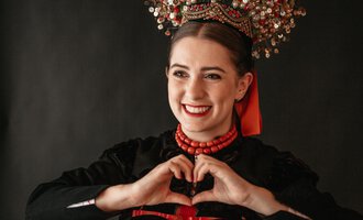 A smiling woman in traditional clothing makes a heart with her hands. Her outfit consists of many colorful beads and she stands against a dark background.