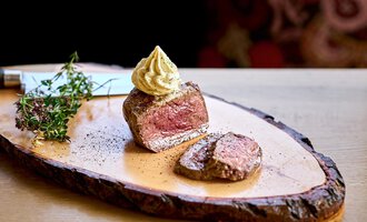 Ein Stück zartes Fleisch auf einem Holzbrett, dekoriert mit Senf und frischen Kräutern. Im Hintergrund sieht man eine stilvolle, gemusterte Textilie. | © Hotel Tradizio | © Annette Sandner