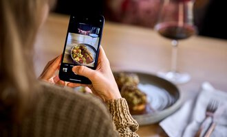 Eine Person macht ein Foto von ihrem Essen mit einem Smartphone. Im Hintergrund ist ein Glas Wein und ein gedeckter Tisch zu sehen. | © Hotel Tradizio | © Annette Sandner