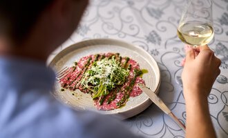 A plate with tender carpaccio, green salad, and parmesan, served with a glass of white wine. In the background, a person can be seen enjoying the dish. | © Hotel Tradizio | © Annette Sandner