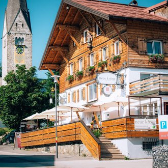 Ein traditionelles, holzverkleidetes Gebäude mit Balkon und Terrassen. Im Hintergrund ist ein Kirchturm und Bäume zu sehen.