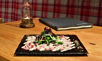 Ein schön angerichteter Teller mit feinem Fleisch und frischen Kräutern. Im Hintergrund sind eine Lampe und ein Kochbuch zu sehen. | © Hotel Neue Krone GmbH | Werbewind GmbH
