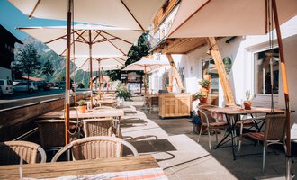 Ein gemütliches Café mit Terrassenstühlen und Sonnenschirmen. Die Straßen sind ruhig und es gibt Pflanzen zur Dekoration.