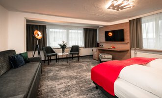 Ein modernes Hotelzimmer mit einem roten Bett und einer gemütlichen Sitzecke. Helle Fenster und stilvolle Möbel schaffen eine einladende Atmosphäre.