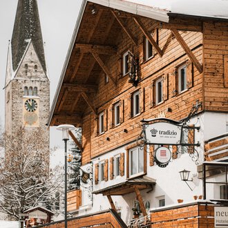 Ein traditionelles Holzhaus im Schnee mit einem Glockenturm im Hintergrund. Die Szene vermittelt eine gemütliche Winteratmosphäre.