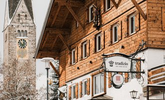 Ein traditionelles Holzhaus im Schnee mit einem Glockenturm im Hintergrund. Die Szene vermittelt eine gemütliche Winteratmosphäre.