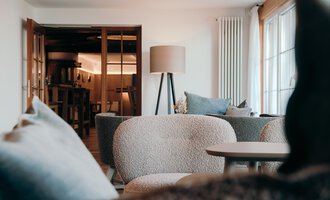 Ein modernes Wohnzimmer mit gemütlichen Sofas und einem Tisch. Im Hintergrund sind Türen und ein Stehlampe zu sehen.