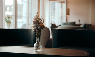 Ein elegantes Wohnzimmer mit einem Holztisch und einem kleinen Glasvase, die mit Trockenblumen gefüllt ist. Im Hintergrund sind bequeme Sitzmöbel und große Fenster zu sehen.