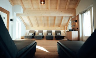 Ein modernes Zimmer mit Holzdecke und Liegen. Es gibt große Fenster, die viel Licht hereinlassen.