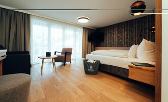 Ein modernes Hotelzimmer mit Holzfußboden und großzügigem Fenster. Die Einrichtung umfasst ein Bett, einen Sessel und einen Tisch.