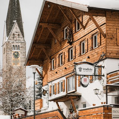 A traditional wooden house in the snow with a bell tower in the background. The scene conveys a cozy winter atmosphere.
