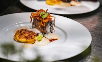 An artistically arranged plate with fried fish, crispy onions, and a side of fried bananas. Accompanied by a creamy sauce and fresh herbs. | © Verwöhn- & Wellnesshotel Walserhof| Frank Simon
