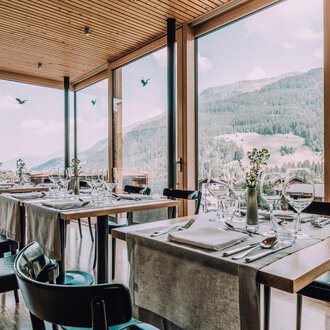 © Naturhotel Chesa Valisa | Heldentheater Ein modernes Restaurant mit großen Fenstern und Blick auf die Berge. Die Tische sind elegant gedeckt und schaffen eine einladende Atmosphäre. | © Naturhotel Chesa Valisa | Heldentheater