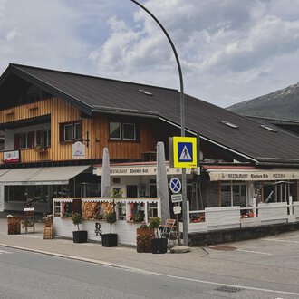 Ein traditionelles alpenländisches Gebäude mit Holzverkleidung und einem Restaurant im Erdgeschoss. Im Vordergrund sind Blumenarrangements und Verkehrssignale zu sehen. | © Riezlern Eck | Sead Sulejmani