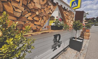 Ein gemütlicher Außenbereich mit einem Holztresen und dekorativen Pflanzen. Im Hintergrund sind Holzstapel und eine Straße sichtbar. | © Riezlern Eck | Sead Sulejmani