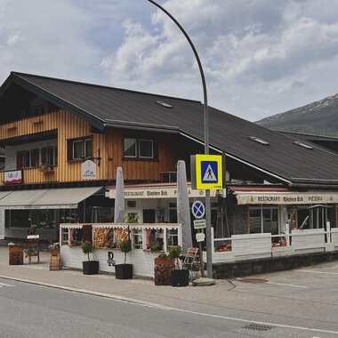 Ein traditionelles alpenländisches Gebäude mit Holzverkleidung und einem Restaurant im Erdgeschoss. Im Vordergrund sind Blumenarrangements und Verkehrssignale zu sehen. | © Riezlern Eck | Sead Sulejmani