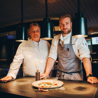 Zwei Köche stehen in einer modernen Küche hinter einem Teller mit elegant angerichtetem Essen. Die Atmosphäre ist einladend und kreativ, mit sanfter Beleuchtung. | © Hotel Birkenhöhe | Stefan Klauser