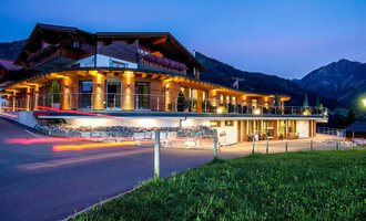 Ein modernes Gebäude im alpinen Stil, beleuchtet von warmem Licht. Im Hintergrund sind beeindruckende Berge zu sehen, während der Himmel in der Dämmerung leuchtet. | © Hotel Birkenhöhe | GA Service Salzburg