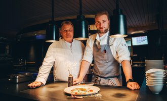 Zwei Köche stehen in einer modernen Küche hinter einem Teller mit elegant angerichtetem Essen. Die Atmosphäre ist einladend und kreativ, mit sanfter Beleuchtung. | © Hotel Birkenhöhe | Stefan Klauser