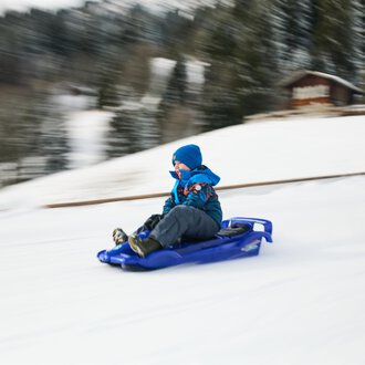 Children are sledding in the snow while adults in warm clothes accompany them. In the background, there is a church and the blue sky is visible. | © Kleinwalsertal Tourismus | Oliver Farys