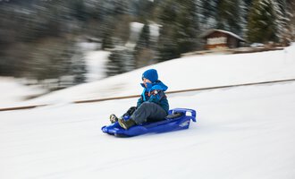 Children are sledding in the snow while adults in warm clothes accompany them. In the background, there is a church and the blue sky is visible. | © Kleinwalsertal Tourismus | Oliver Farys