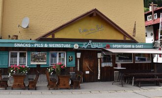 A cozy inn with a colorful sign and blooming flowers on the terrace. The façade is in warm colors and conveys an inviting atmosphere. | © s‘Älpele | Marco Mitterer