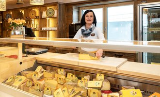 Eine Käseverkäuferin steht hinter einer Theke voller Käsesorten. Der Laden hat ein gemütliches Ambiente mit Holzregalen und natürlichen Lichtquellen. | © Schönegger Käse-Alm