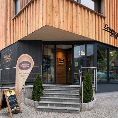 Ein modernes Gebäude mit Holzfassade und großen Fenstern. Vor dem Eingang steht ein Schild mit einer Einladung. | © Schönegger Käse-Alm