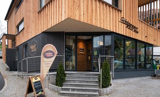 A modern building with a wooden facade and large windows. In front of the entrance stands a sign with an invitation. | © Schönegger Käse-Alm