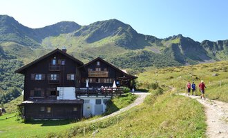 Eine gemütliche Berghütte mit einer wunderschönen Landschaft im Hintergrund. Wanderer genießen den sonnigen Tag auf dem Weg zur Hütte. | © Schwarzwasserhütte | Dominik Müller