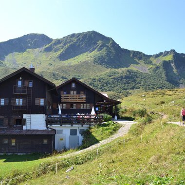 Eine gemütliche Berghütte mit einer wunderschönen Landschaft im Hintergrund. Wanderer genießen den sonnigen Tag auf dem Weg zur Hütte. | © Schwarzwasserhütte | Dominik Müller