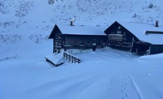 Eine ruhige Winterlandschaft mit einem Chalet, das von einer dicken Schneedecke umgeben ist. Die Berge im Hintergrund sind ebenfalls schneebedeckt. | © Schwarzwasserhütte | Tine Müller