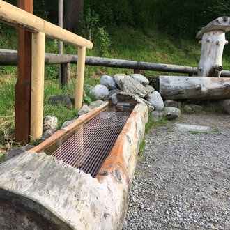Ein Holzbrunnen aus Baumstämmen steht neben einer Stein- und Grasfläche. Dahinter sind weitere Holzstrukturen zu sehen, umgeben von Bäumen. | © Kleinwalsertal Tourismus