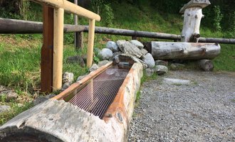 Ein Holzbrunnen aus Baumstämmen steht neben einer Stein- und Grasfläche. Dahinter sind weitere Holzstrukturen zu sehen, umgeben von Bäumen. | © Kleinwalsertal Tourismus