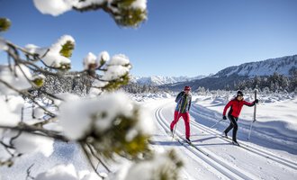 Two people are skiing on a snow-covered track. In the background, there are snow-capped mountains and a clear blue sky. | © Kleinwalsertal Tourismus | Dominik Berchtold