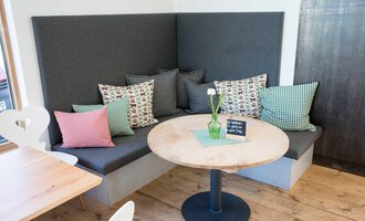 A cozy café area with a padded bench and colorful cushions. On the table, there is a small vase with a flower. | © Kleinwalsertal Tourismus | Frank Drechsel