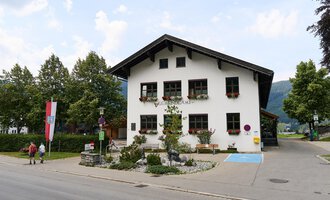 A charming, traditional building with a hipped roof. It is surrounded by trees and has blooming window boxes. | © Kleinwalsertal Tourismus | Oliver Farys