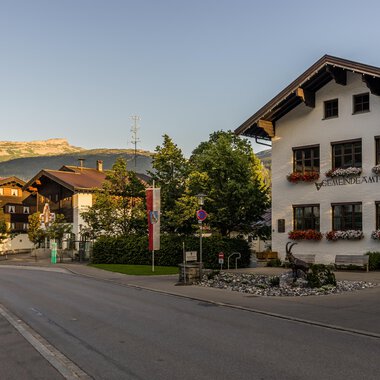 A quiet street with typical Bavarian houses and blooming windows. In the background, mountains are visible and the sky is clear. | © Kleinwalsertal Tourismus | Steffen Berschin