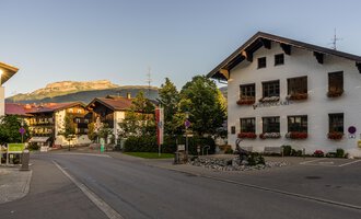 A quiet street with typical Bavarian houses and blooming windows. In the background, mountains are visible and the sky is clear. | © Kleinwalsertal Tourismus | Steffen Berschin