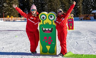 Zwei fröhliche Menschen in roten Anzügen stehen neben einem bunten Monster-Schild im Schnee. Im Hintergrund sind Bäume und eine verschneite Landschaft zu sehen.