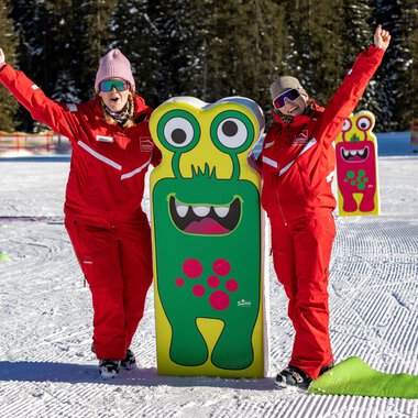 Zwei fröhliche Menschen in roten Anzügen stehen neben einem bunten Monster-Schild im Schnee. Im Hintergrund sind Bäume und eine verschneite Landschaft zu sehen.
