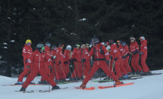 Eine Gruppe von Skifahrern in roten Anzügen steht in einem verschneiten Skigebiet. Die Bäume im Hintergrund sorgen für eine winterliche Atmosphäre.