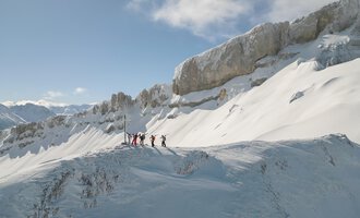 Eine Gruppe von Menschen steht auf einem schneebedeckten Berggipfel. Im Hintergrund sind majestätische Felsen und ein klarer blauer Himmel zu sehen. | © Skischule Hirschegg GmbH
